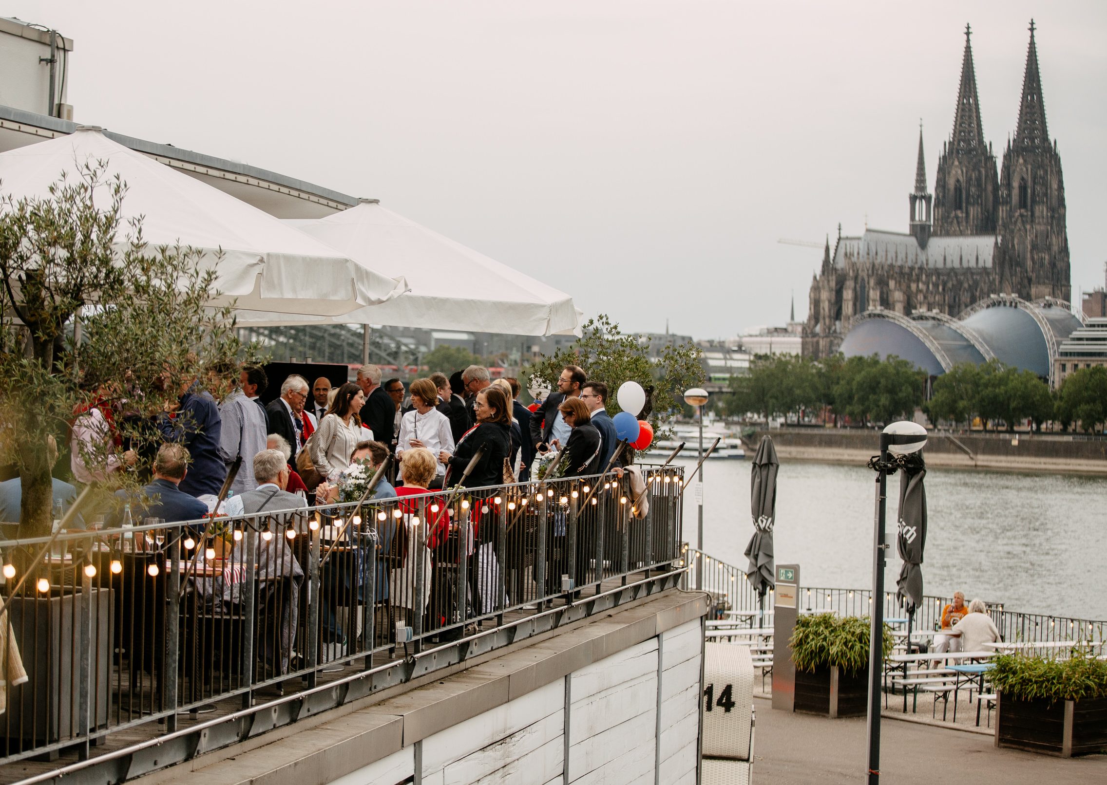 Auf der linken Bildhälfte steht eine Menschenmenge auf einer Terrasse. In der rechten Bildhälfte sieht man den Rhein und den Kölner Dom.