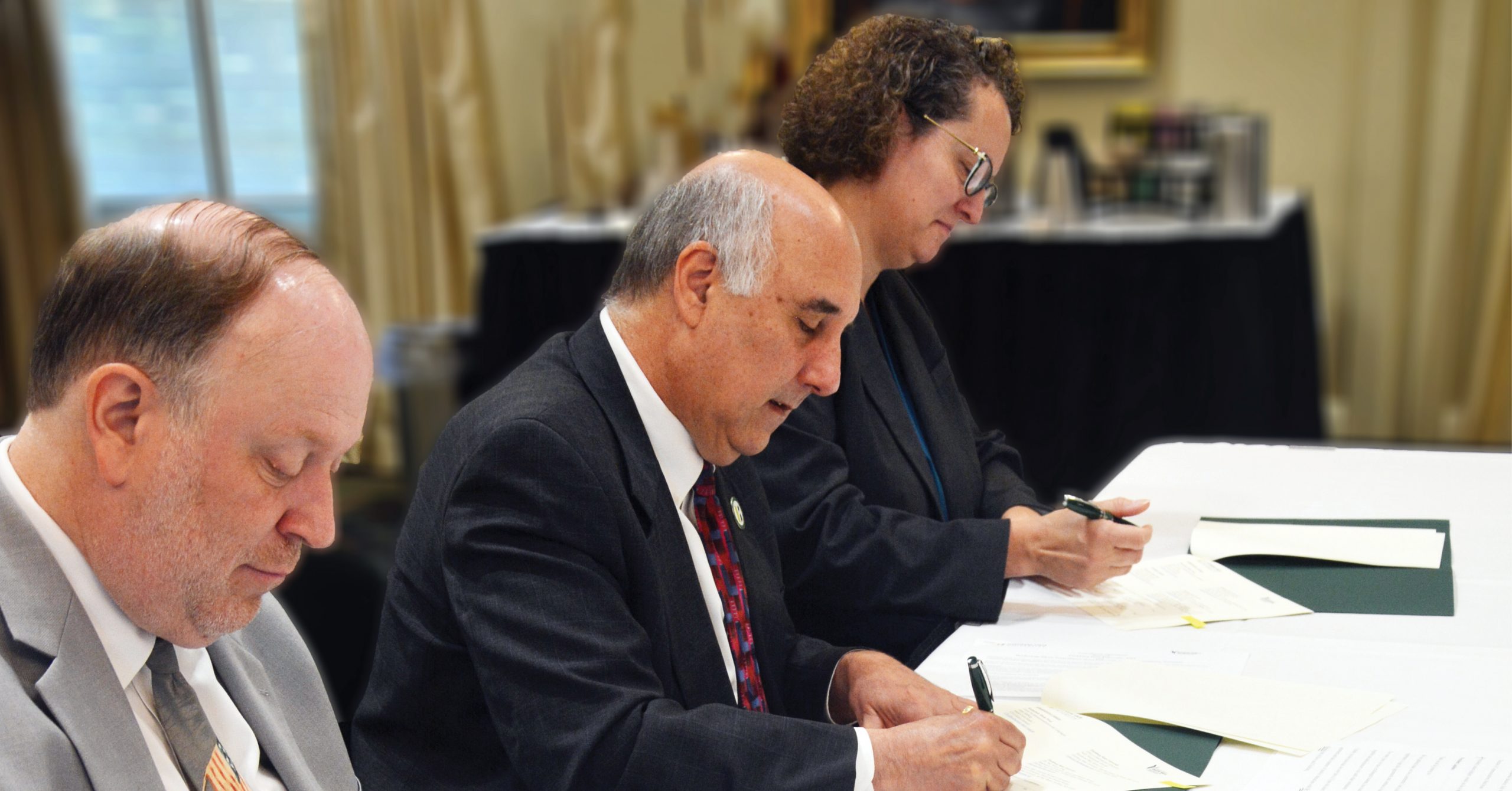 Drei Personen sitzen nebeneinander an einem Tisch und unterzeichnen Papiere